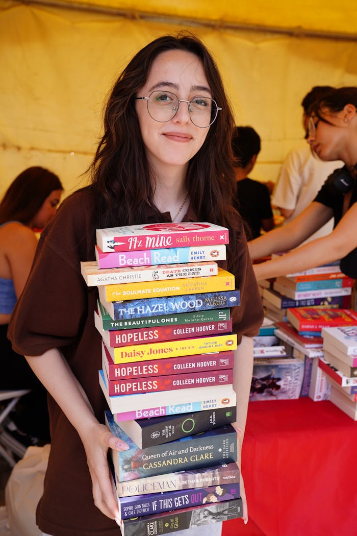 gallery-3 A young woman in glasses smiles while holding a large stack of popular books at an indoor event.